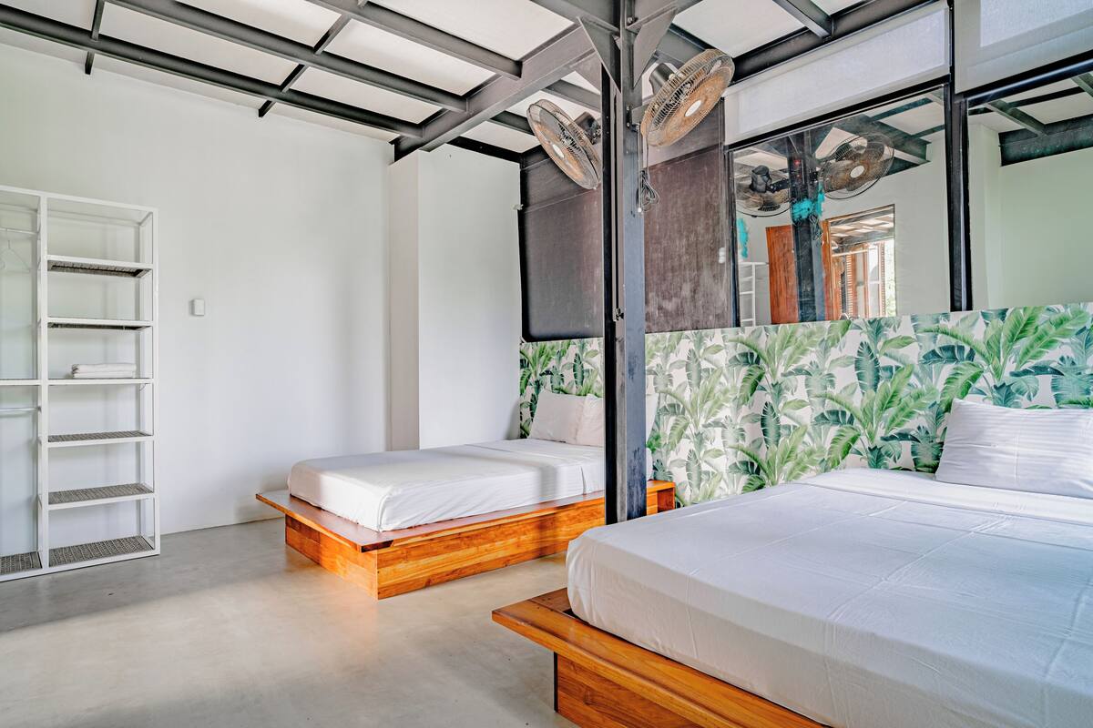 A modern bedroom with two beds, a white shelving unit, and a large window with a view of greenery.