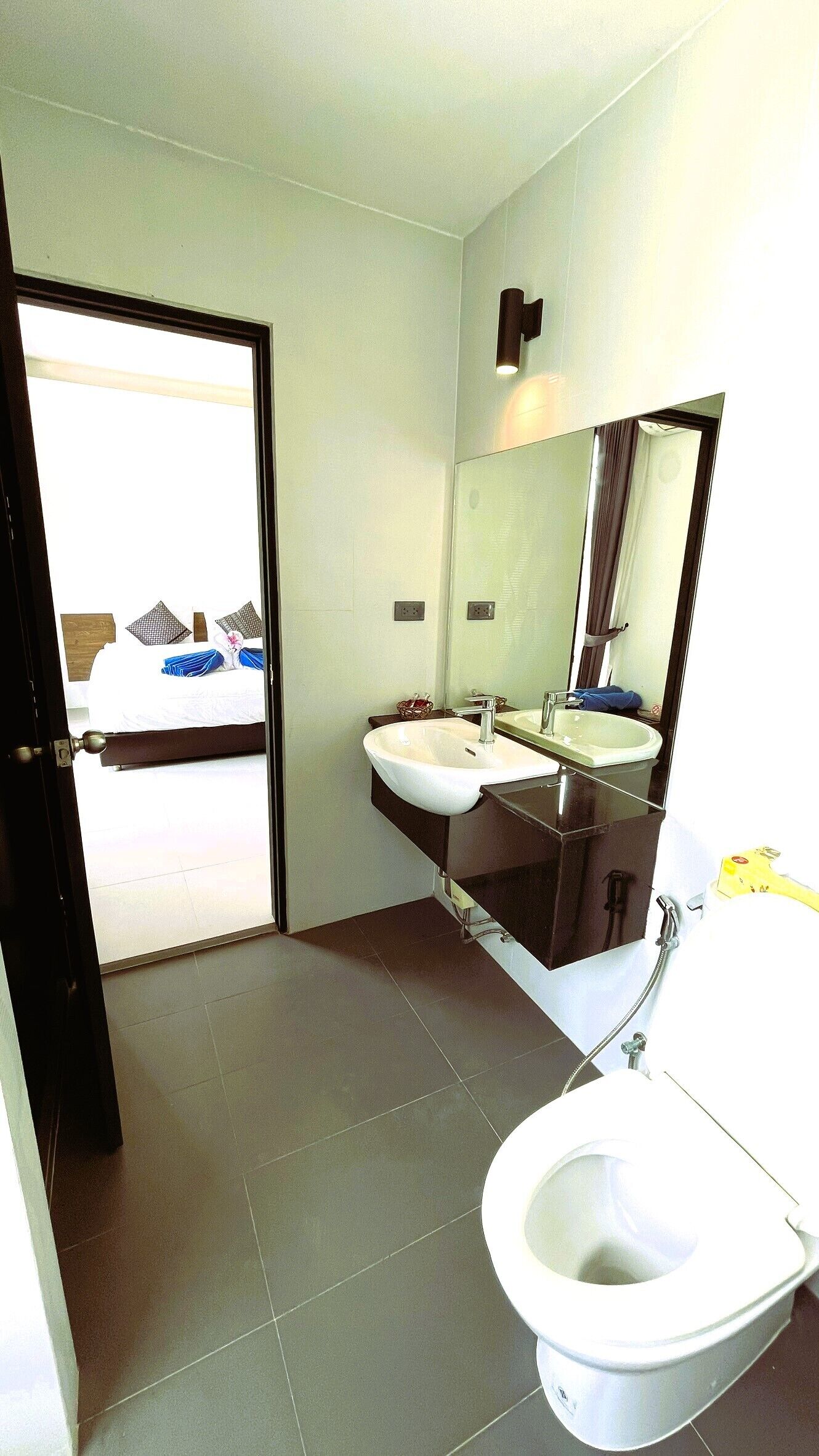 Casa de banho de hotel com sanita, duas lavatórios, um espelho grande e uma cama visível por uma porta aberta.