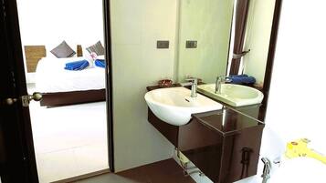 Casa de banho de hotel com sanita, duas lavatórios, um espelho grande e uma cama visível por uma porta aberta.