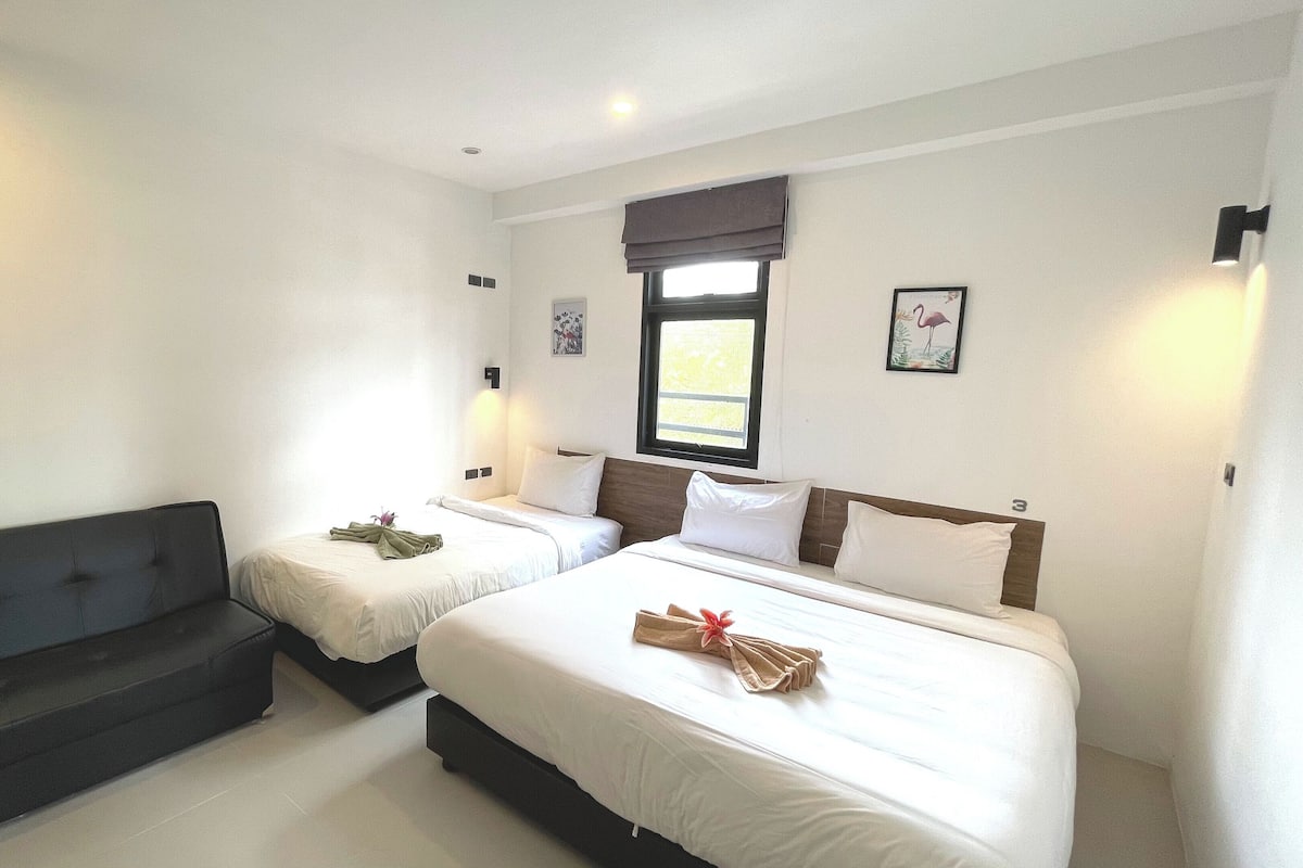 A hotel room with two single beds, a sofa, a window with a view, and a small table with a flower.