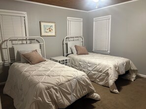 Une chambre avec deux lits simples, une petite table de chevet, un tableau encadré au mur et un ventilateur de plafond.