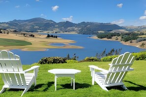 Deux chaises Adirondack blanches et une petite table blanche sont placées sur une pelouse, offrant une vue sur un lac avec des montagnes en arrière-plan.