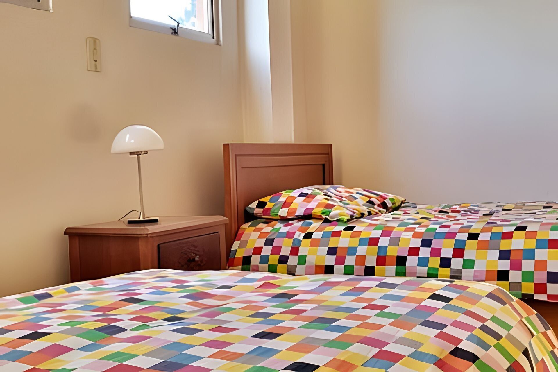 A hotel room with two beds, each with a colorful checkered bedspread. There is a wooden nightstand with a lamp and a small drawer.