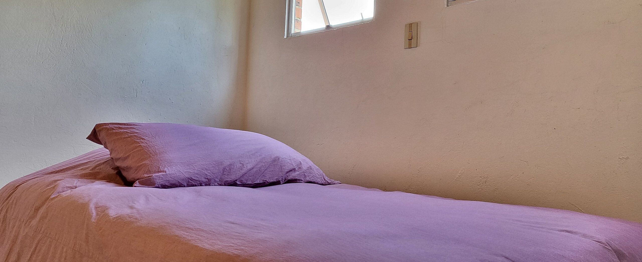 A single bed with a purple bedspread and pillows in a room with plain walls and a small window.