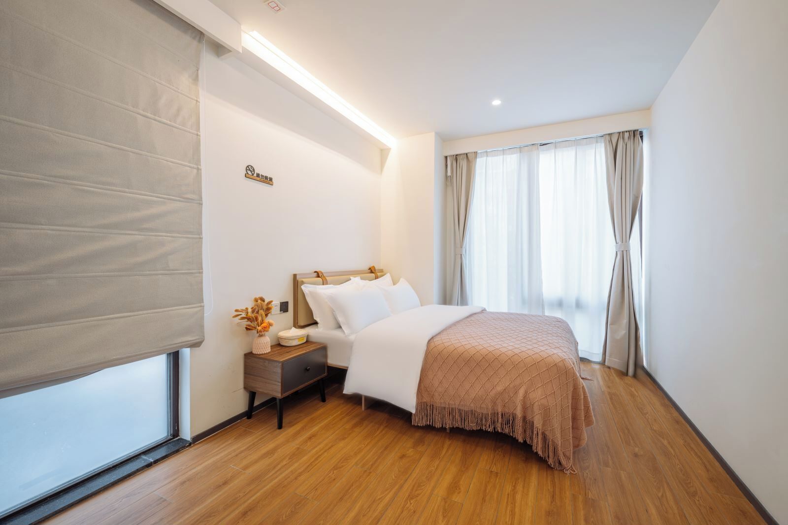 A hotel room with a bed, a nightstand, a lamp, a window with curtains, and a sign on the wall.