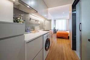 Una camera d'albergo compatta con angolo cottura, lavatrice, un letto con copriletto arancione e vista sulla città dalla finestra.