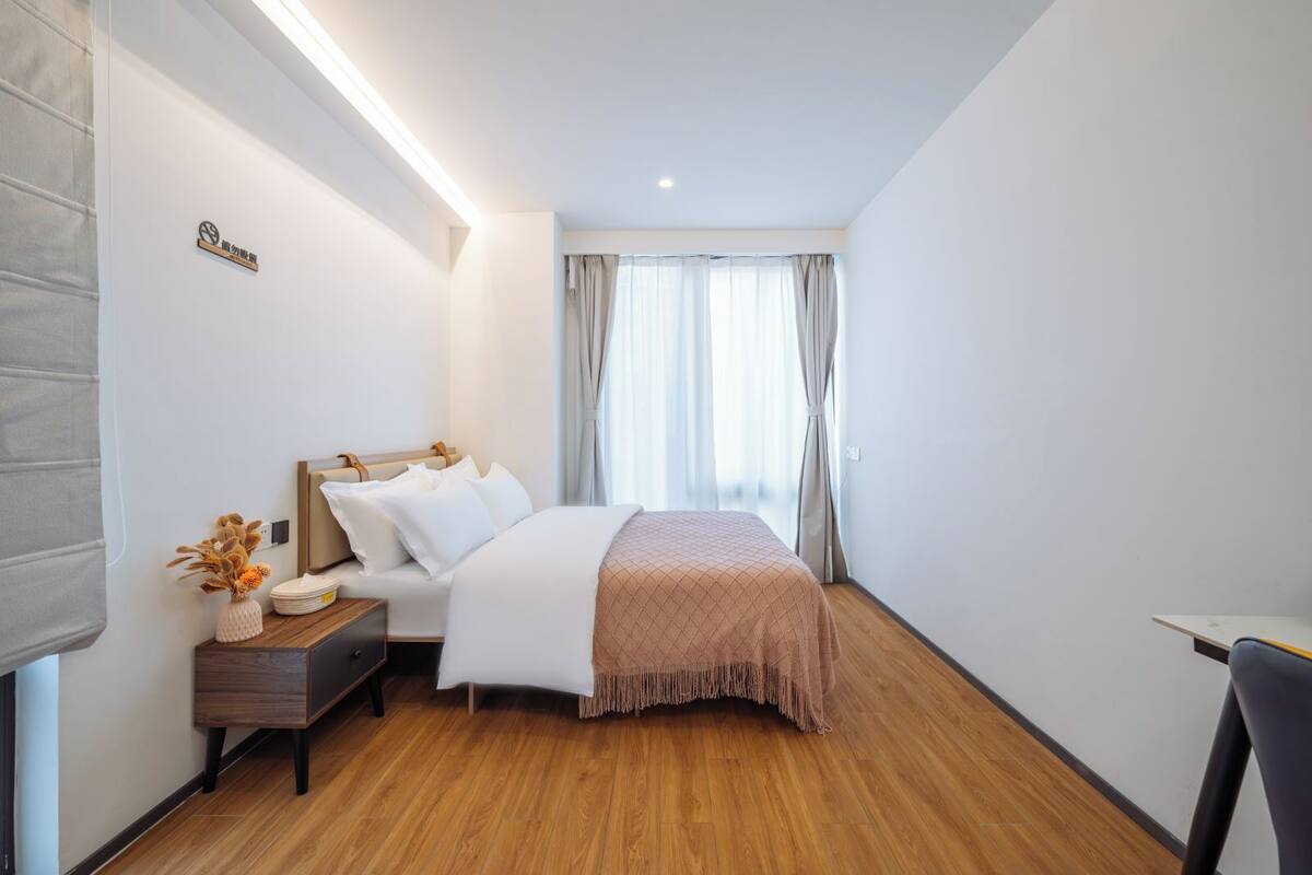 A modern hotel room with a wooden floor, a bed with white bedding and a pink throw, a bedside table with a vase of flowers, and a window with sheer curtains.