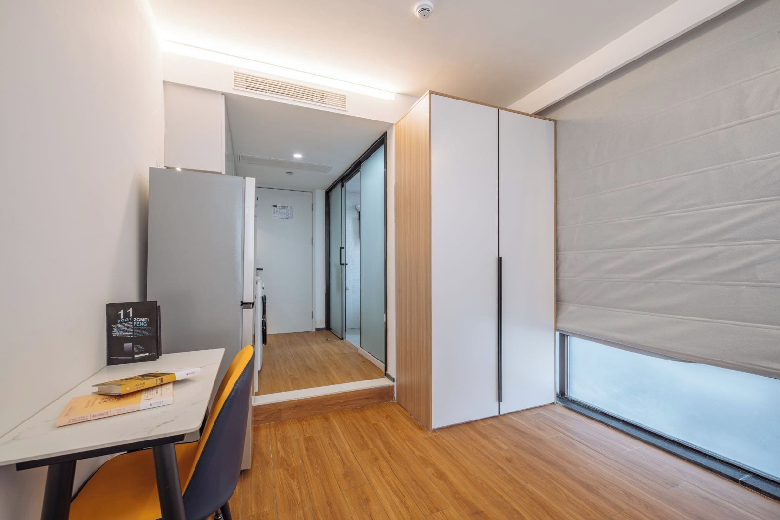 A modern hotel room with a wooden floor, a desk with a chair, a bed with a headboard, and a large window with blinds.