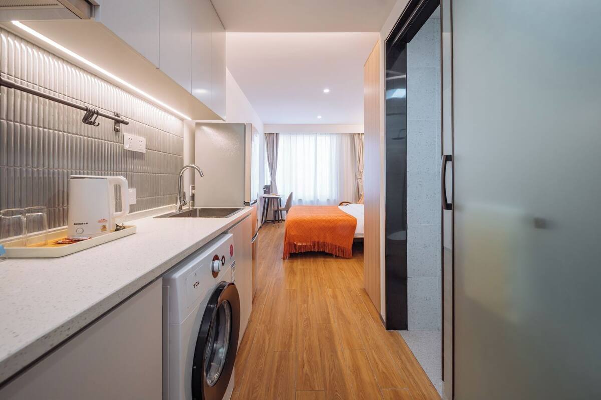 A compact hotel room with a kitchenette, a washing machine, a bed with an orange bedspread, and a view of the city through the window.