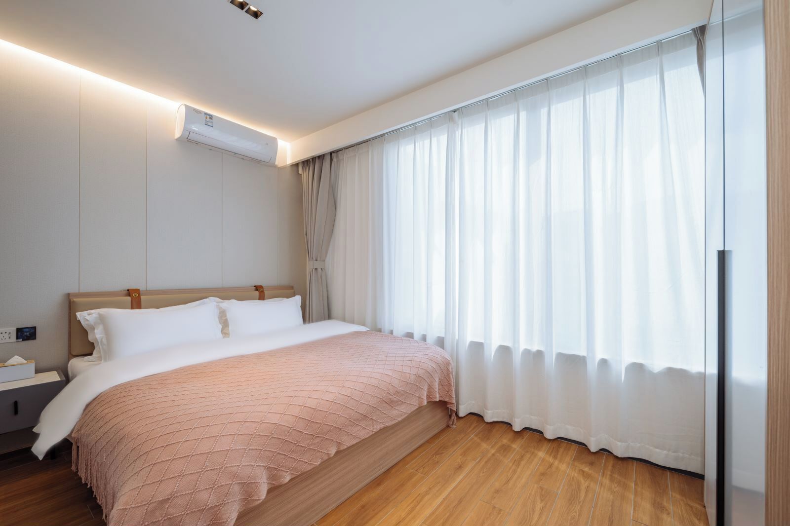 Quarto de hotel com cama, mesa de cabeceira, ar condicionado e uma janela grande com cortinas.