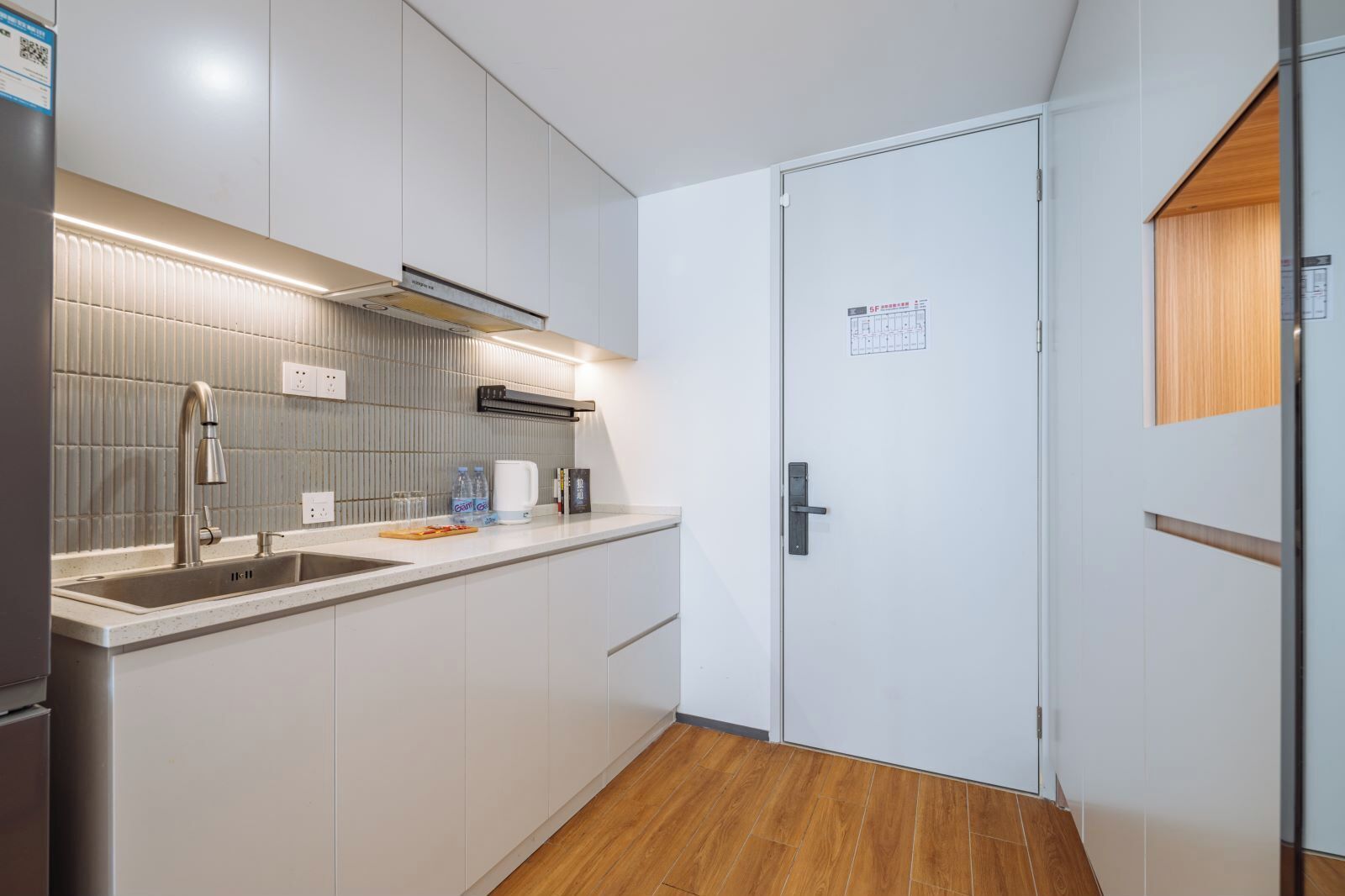 A modern kitchen with a built-in sink, stainless steel faucet, and a microwave. There is a wooden floor and a white door with a small window.