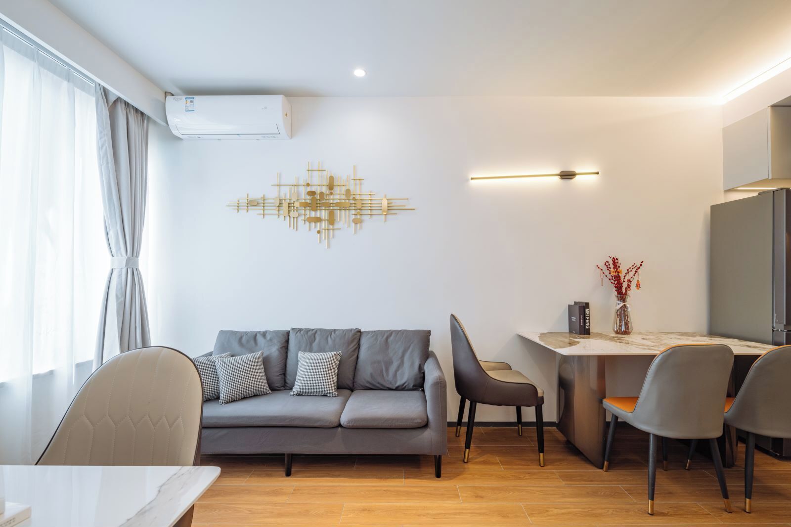 Una sala de estar moderna con un sofá gris, una mesa de comedor y sillas. Hay un aire acondicionado instalado en la pared y un accesorio de iluminación decorativo.