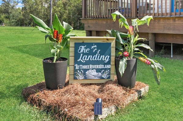 A wooden sign with the text "The Landing at Three Rivers Island" surrounded by potted plants on a grassy area.