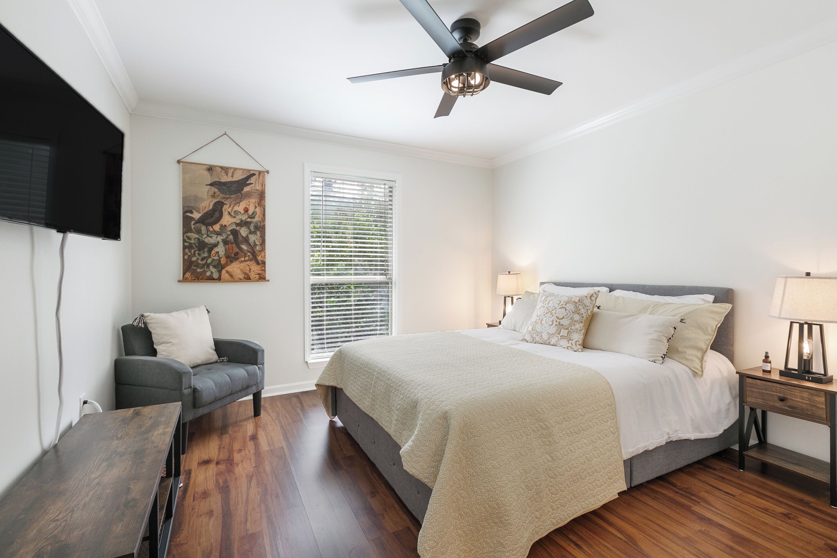 A bedroom with a bed, a chair, a table, a ceiling fan, a window with blinds, and a wall-mounted TV.