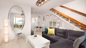 Una sala de estar moderna con un sofá gris, una mesa de centro blanca y un espejo grande. Una escalera con vigas de madera conduce a un nivel superior.