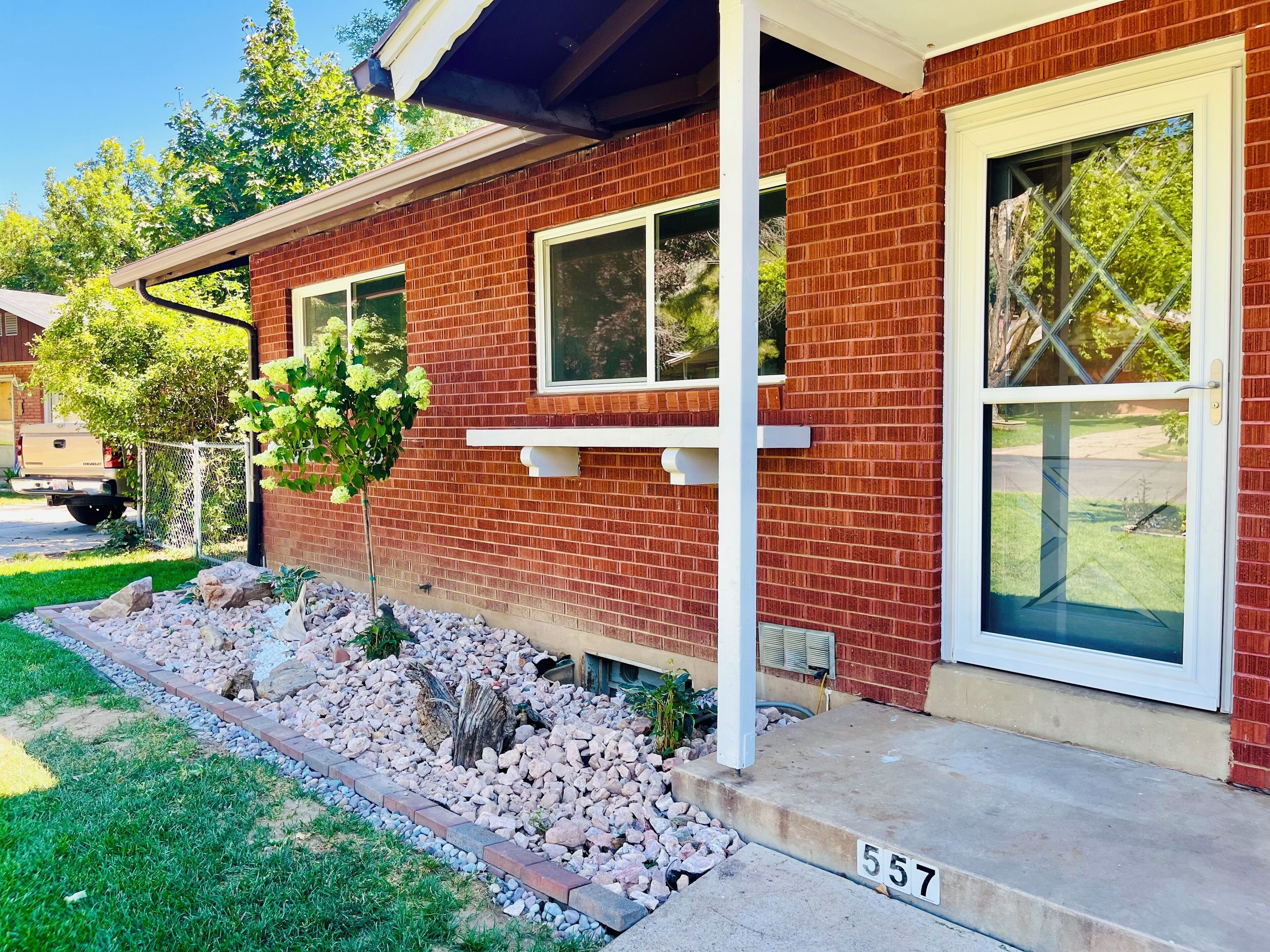 A red brick house with a number '557' on the front steps, a small garden with rocks and plants, and a hanging flower basket.