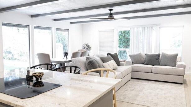 Une cuisine moderne avec un plan de travail en marbre, un coin repas avec une table et des chaises, un canapé beige et de grandes fenêtres offrant une vue sur la verdure.