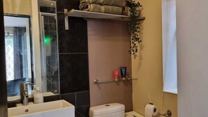 Un bagno con doccia, servizi igienici e lavandino. C'è un portascopino sopra la doccia e uno scaffale con articoli da toilette.