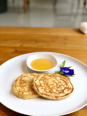 Two pancakes on a white plate with a small bowl of syrup and a blue flower garnish.
