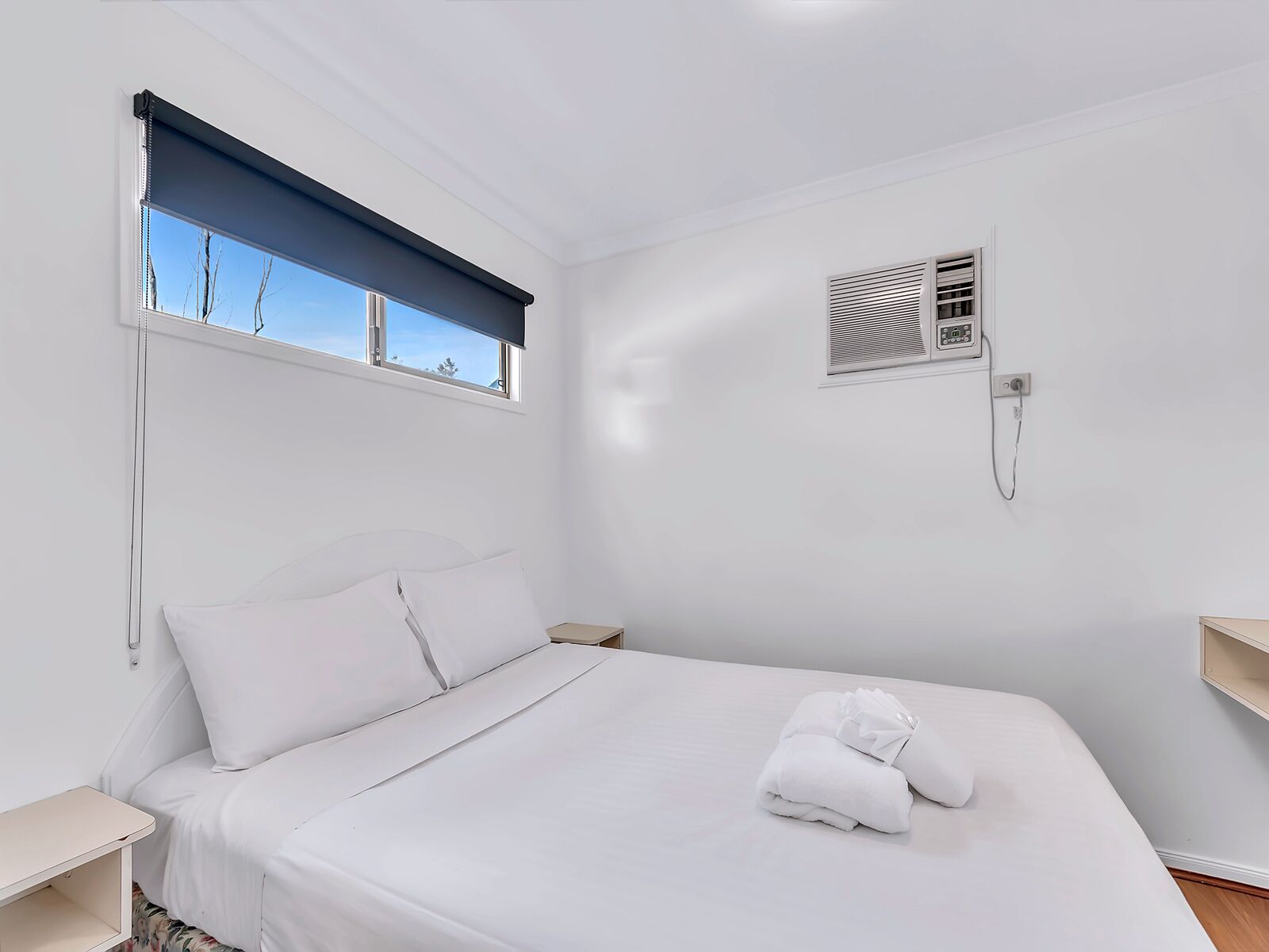Una cama bien hecha con sábanas blancas, una ventana con persiana azul, un aire acondicionado instalado en la pared y una mesita de noche.
