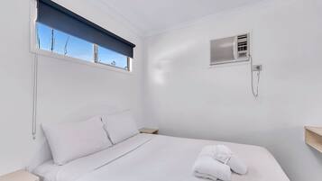 Una cama bien hecha con sábanas blancas, una ventana con persiana azul, un aire acondicionado instalado en la pared y una mesita de noche.
