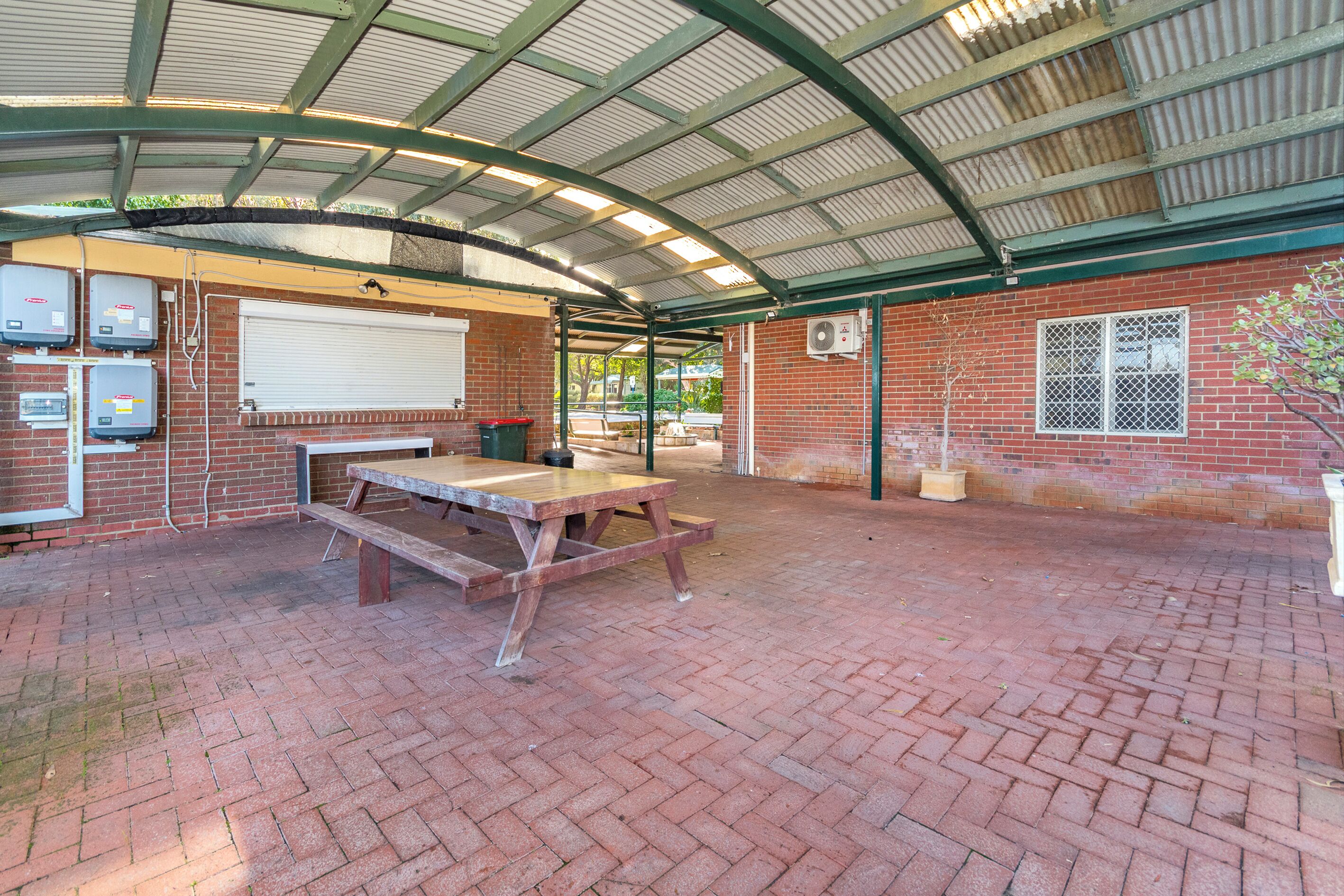 A covered outdoor area with a picnic table, brick flooring, and a metal roof structure.