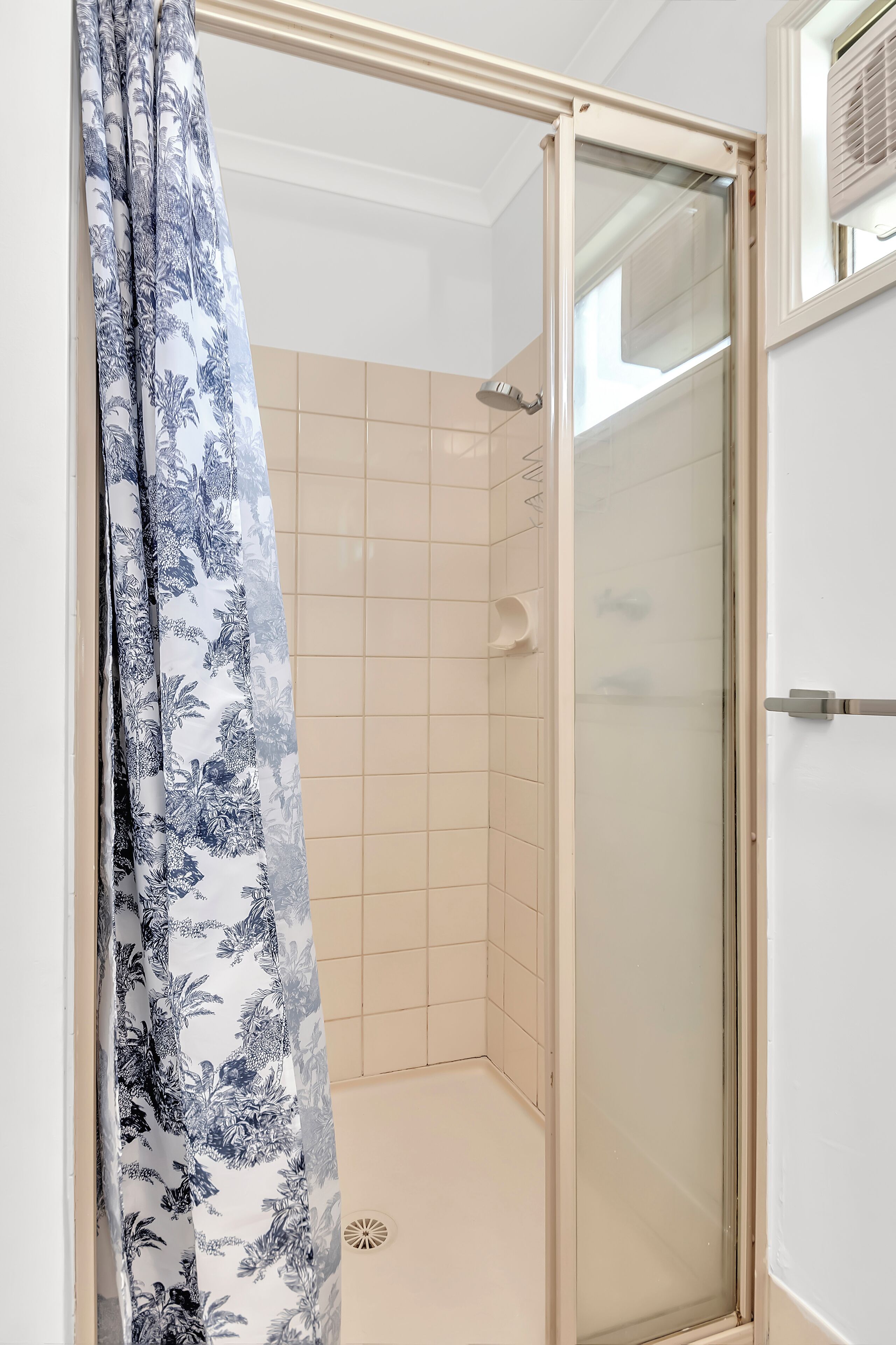 A shower area with a glass door, beige tiles, and a floral curtain.
