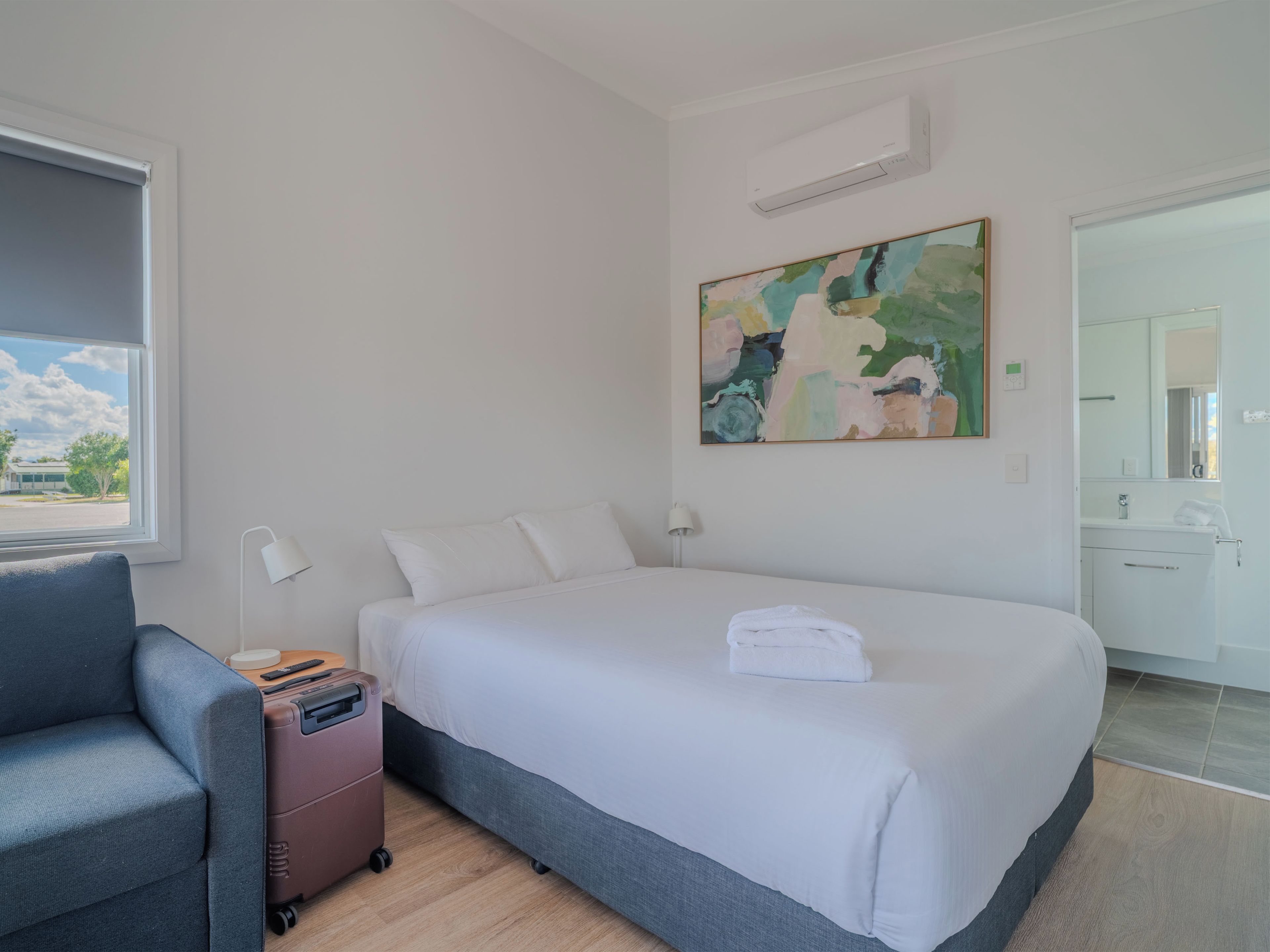 Habitación de hotel con cama, escritorio, silla, maleta, un cuadro y baño visible a través de una puerta abierta.