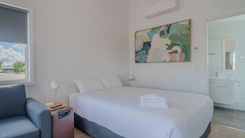 Habitación de hotel con cama, escritorio, silla, maleta, un cuadro y baño visible a través de una puerta abierta.