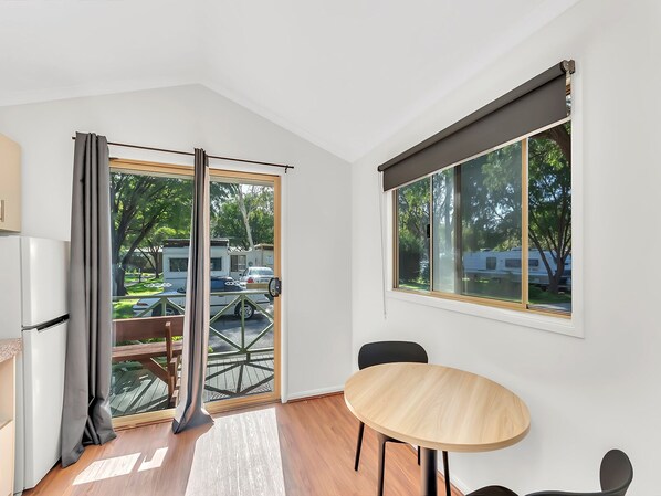 A modern room with a dining table, a refrigerator, and a large window with a view of a parking area and trees.