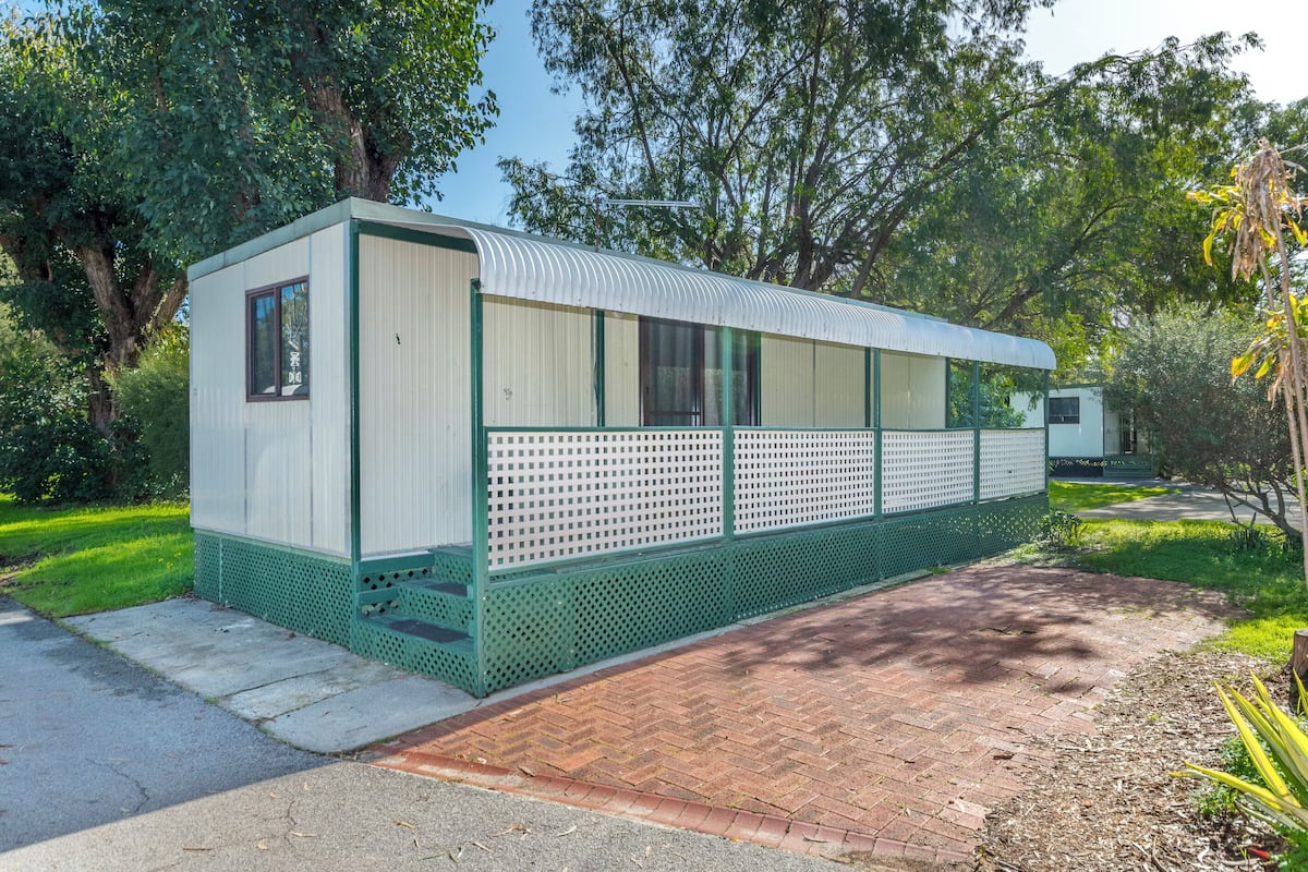 A small, single-story mobile home with a green exterior and white walls, situated on a paved area with a brick pathway leading to it.