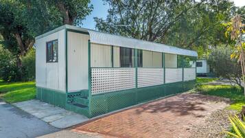 A small, single-story mobile home with a green exterior and white walls, situated on a paved area with a brick pathway leading to it.