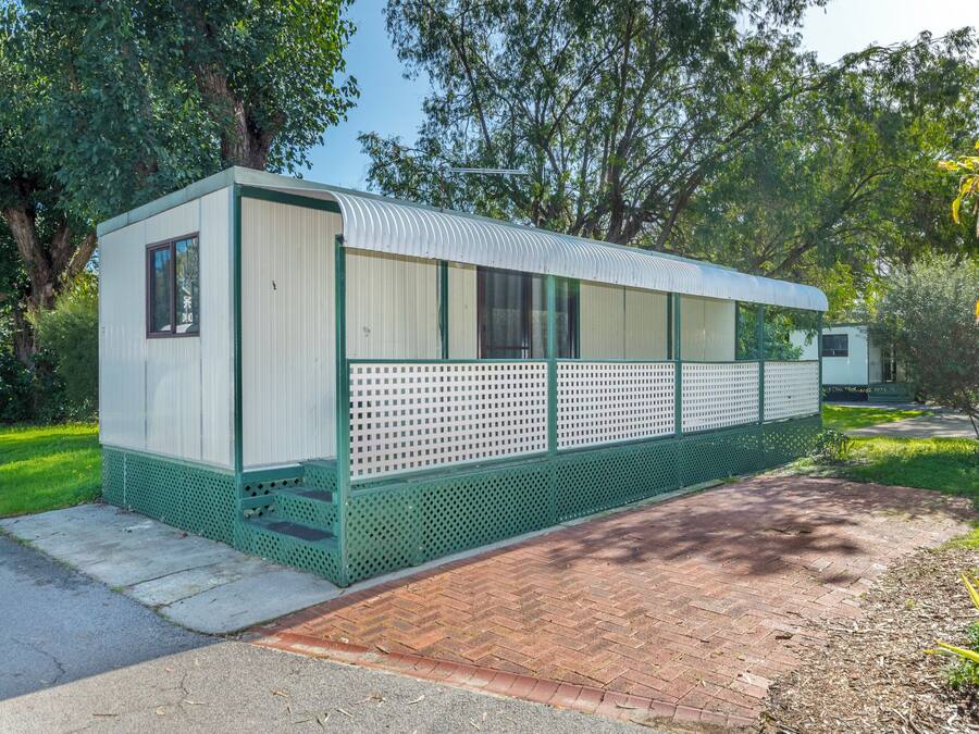 A small, single-story mobile home with a green exterior and white walls, situated on a paved area with a brick pathway leading to it.
