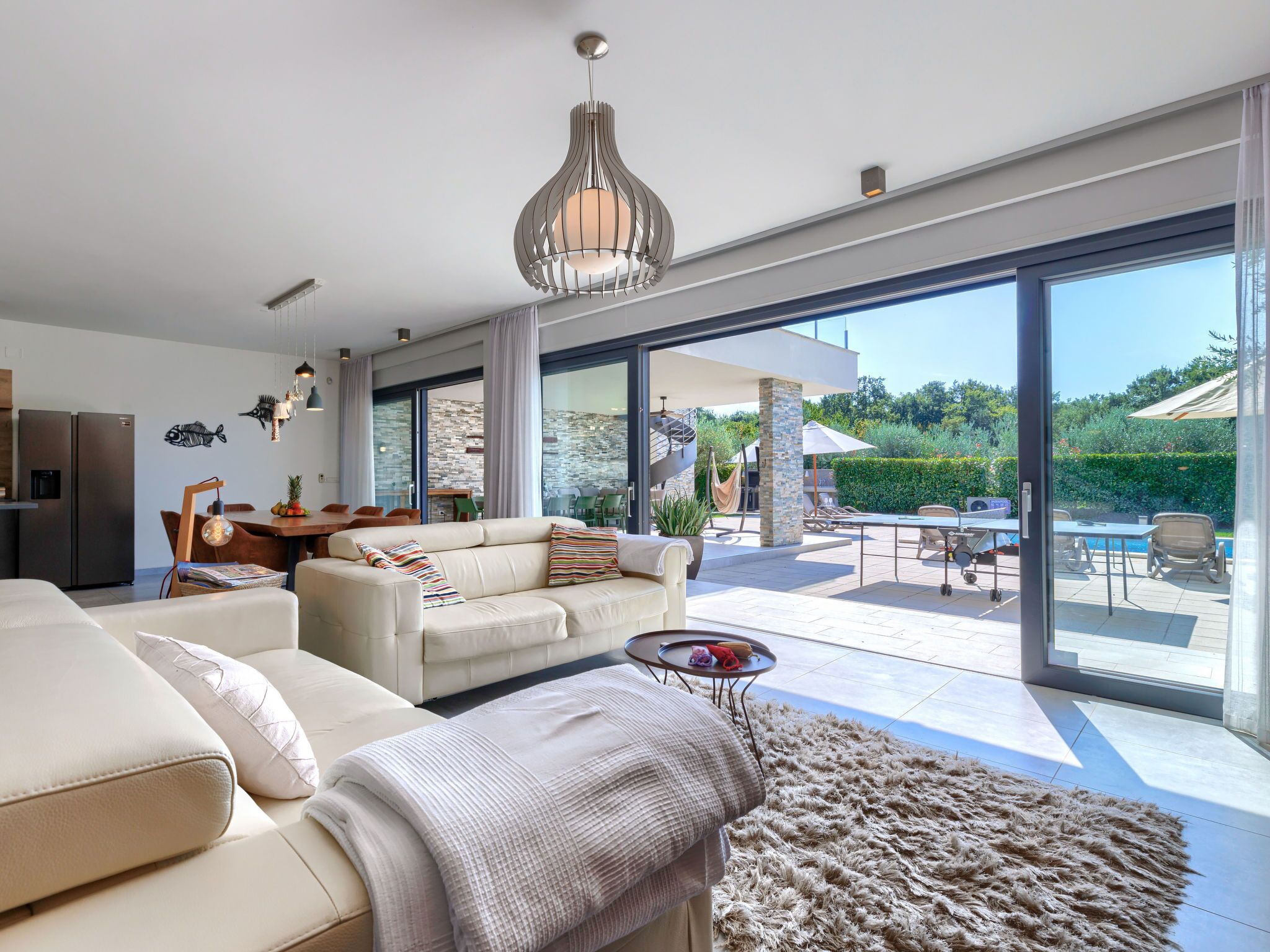 A modern living room with a sofa, a coffee table, and a rug. There is a dining area with a table and chairs. The room has large windows and a view of the outdoors.