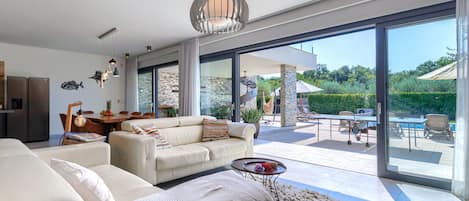 A modern living room with a sofa, a coffee table, and a rug. There is a dining area with a table and chairs. The room has large windows and a view of the outdoors.