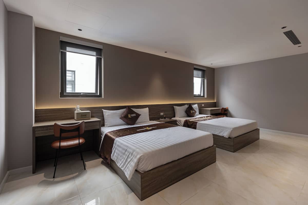 Une chambre d’hôtel moderne, dotée d’un grand lit, d’un bureau et d’une chaise. Deux fenêtres laissent entrer la lumière naturelle.
