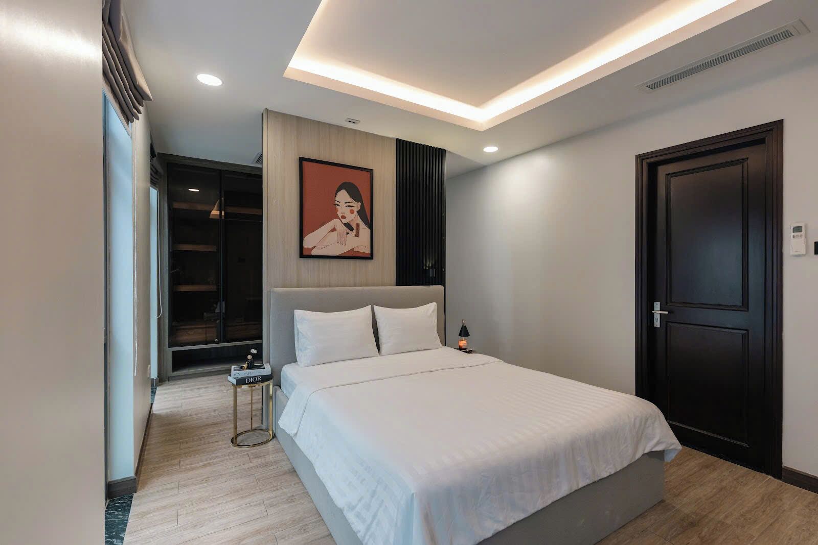 A modern hotel bedroom with a large bed, a bedside table with a lamp, a wardrobe, and a door.