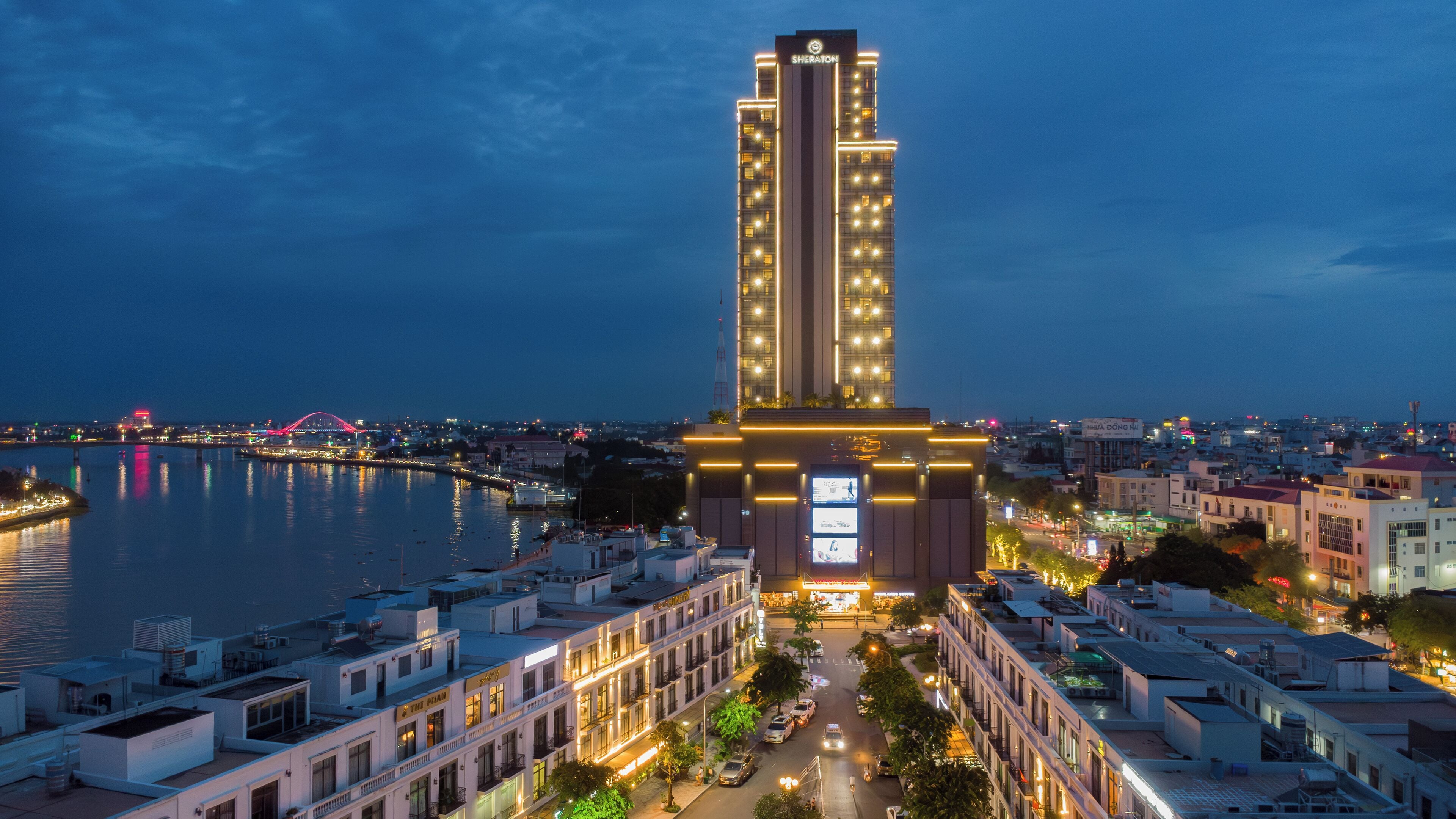 A high-rise hotel with a prominent vertical design, located near a waterfront with a bridge in the background.
