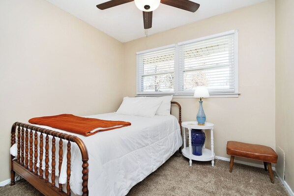 A bedroom with a wooden bed, white bedding, a ceiling fan, a window with blinds, a bedside table with a lamp, and a bench.