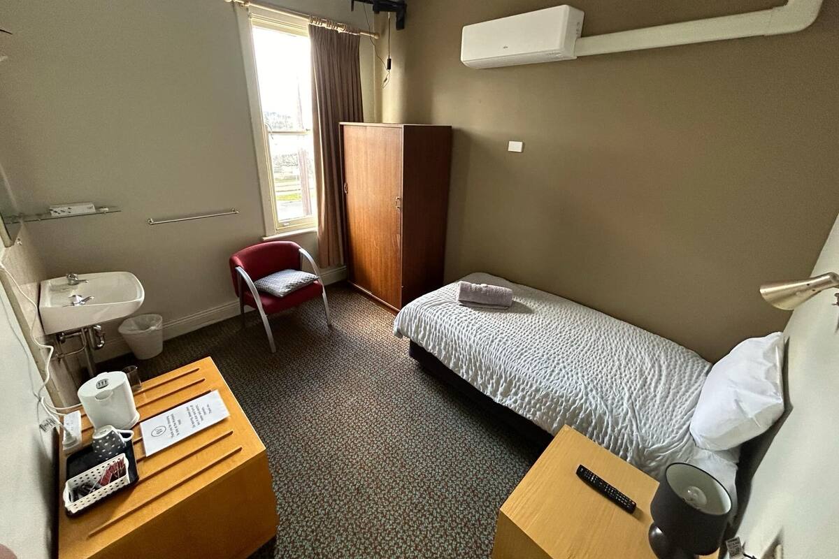 Habitación de hotel con una cama individual, un escritorio con computadora, una silla, un armario, una ventana con cortinas, un lavamanos y un pequeño bote de basura.