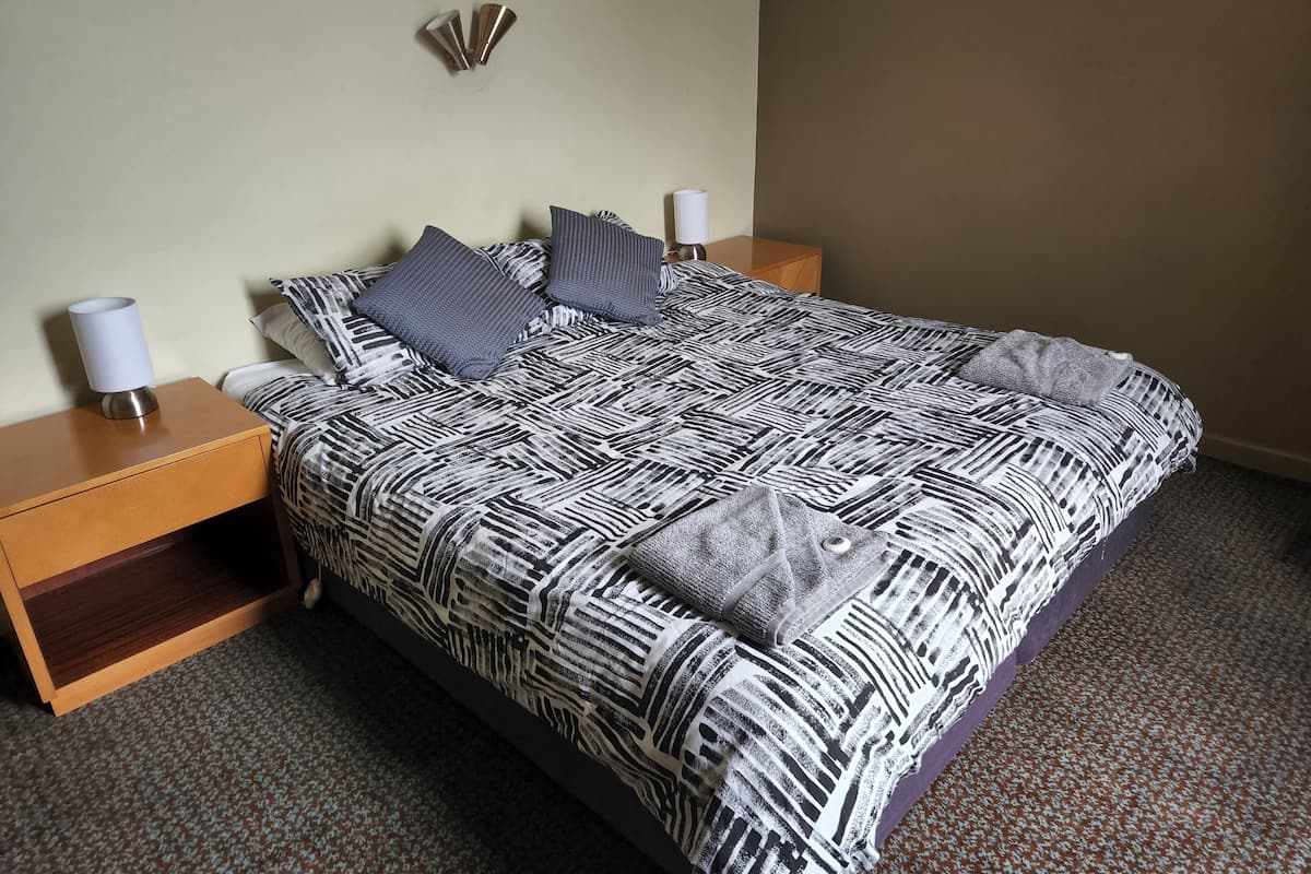 Una cama bien hecha con una colcha estampada, dos almohadas y una lámpara de noche sobre una mesita de madera.