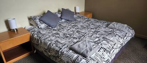 Una cama bien hecha con una colcha estampada, dos almohadas y una lámpara de noche sobre una mesita de madera.