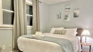 A neatly made bed with a white comforter and a folded towel, a bedside lamp, and a small nightstand with a lamp. - Mid Century Modern 3-bedroom 2-bath house with WiFi, AC, W backyard (Redford Charter Township)