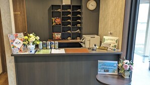 Zona de recepción de un hotel con un mostrador de madera, un reloj, una estantería con artículos de cortesía y un ventanal con vistas.