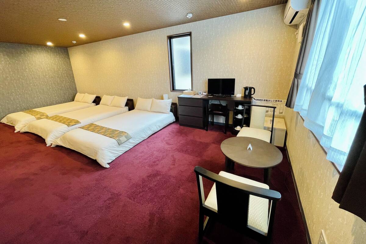 Une chambre d’hôtel avec deux lits, un bureau avec un ordinateur, une chaise, une petite table et une fenêtre avec des rideaux.