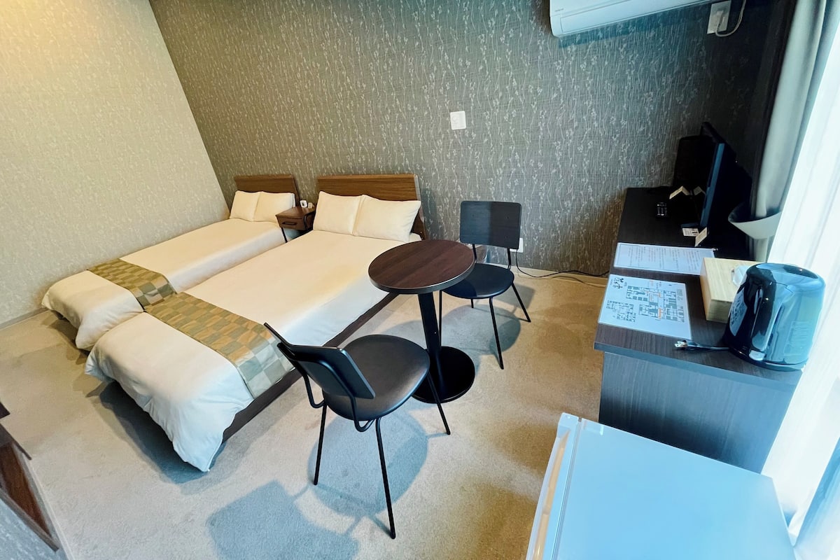 Une chambre d’hôtel avec deux lits simples, une petite table avec deux chaises, un bureau avec un ordinateur et un climatiseur fixé au mur.