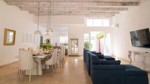Uma sala de estar ampla com mesa de jantar, cadeiras, sofá, luminárias pendentes e uma TV.