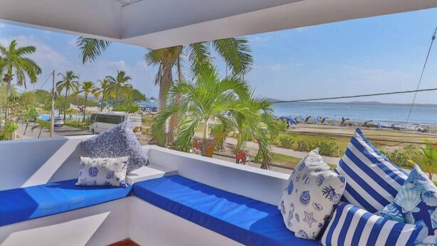 Varanda com almofadas azuis, vista para a praia, palmeiras e céu claro.
