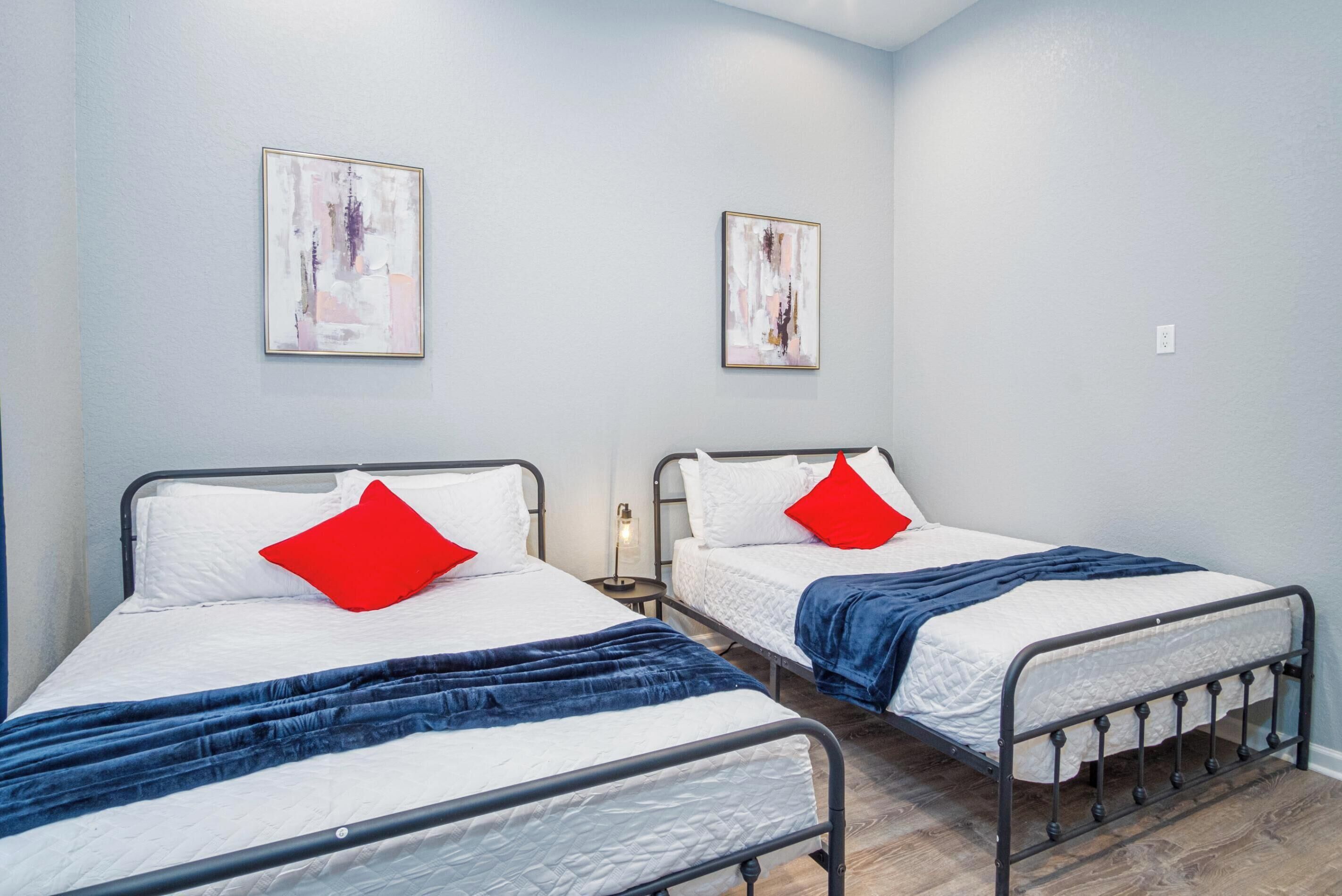 A room with two single beds, each with a metal frame, white bedding, and red pillows. There are abstract paintings on the walls and a small table with a lamp between the beds.