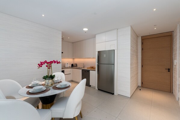 A modern kitchen with white cabinetry, a stainless steel refrigerator, and a dining area with white chairs and a table set for two.